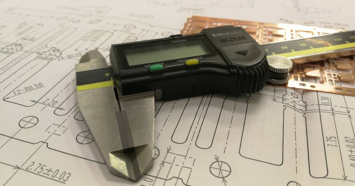 Metal digital caliper with a plastic display placed on a large sheet of paper with multiple numbers and measurements.