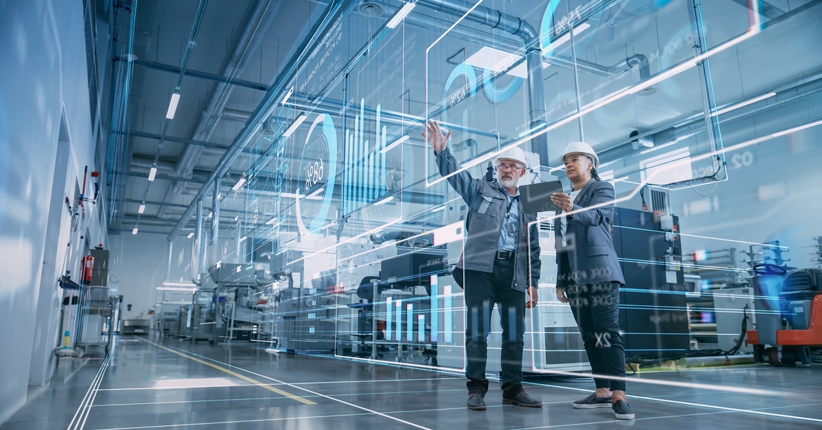 Two people in white hard hats stand together looking at a digital display of different charts and data.