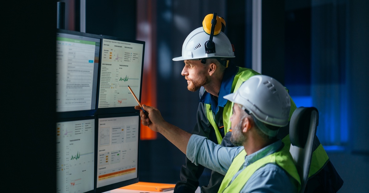 Two workers wearing hi-vis vests and hard hats look at a set of computer screens together. The screens show text and charts.