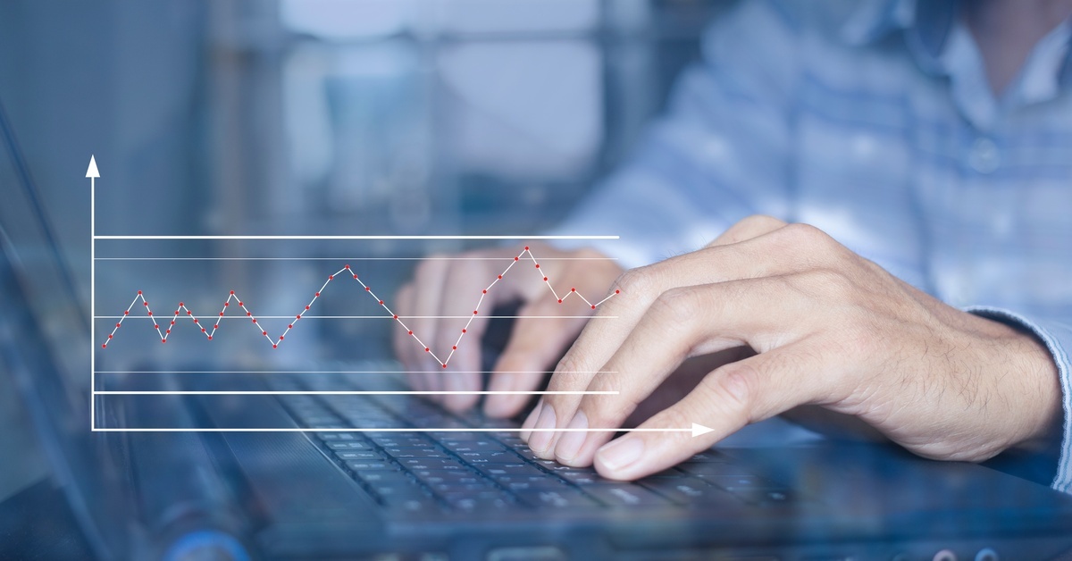 A close-up of a person in business attire typing on a laptop. There is a line chart superimposed over the keyboard.