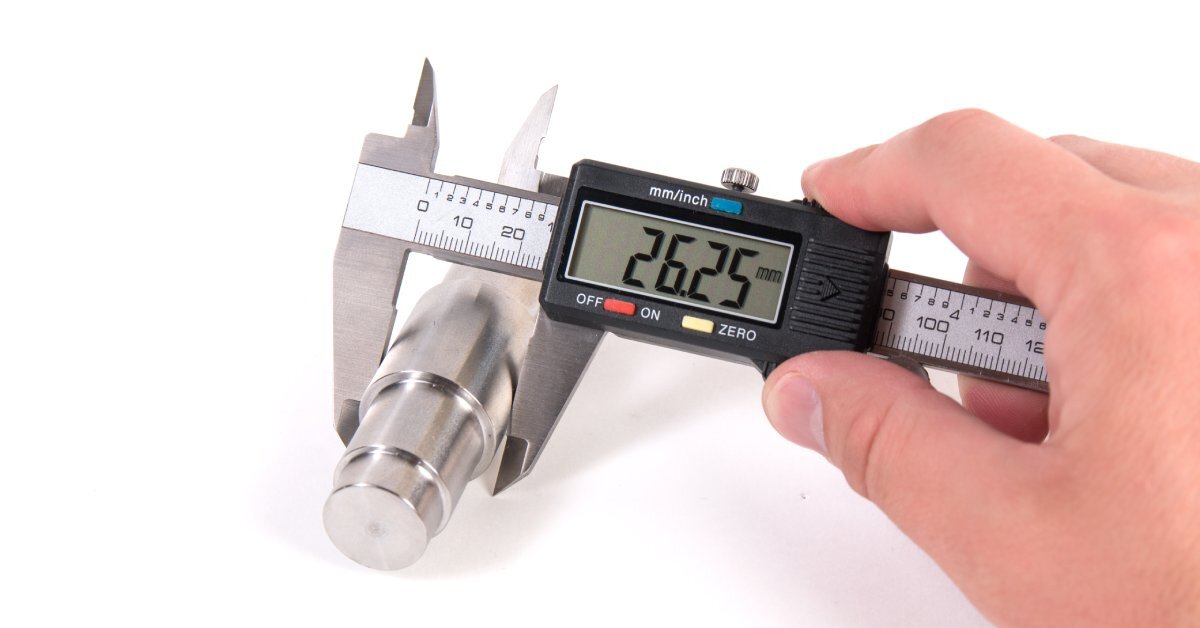 A person holding a digital caliper measuring a cylindrical metal part. The display reads "26.25 mm."