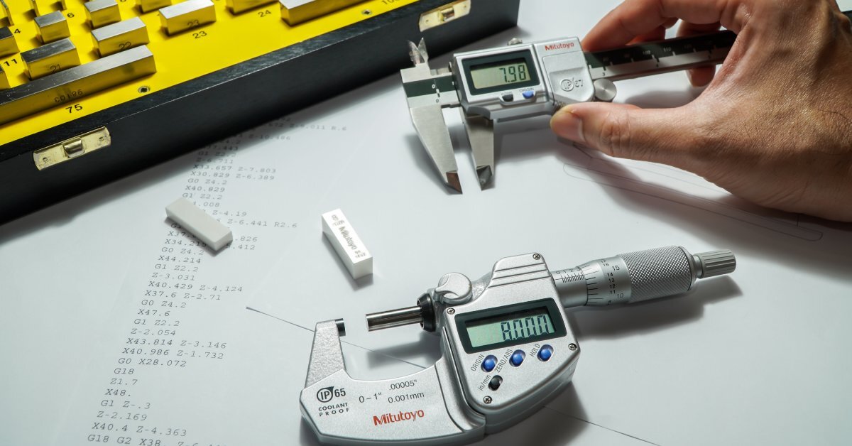 A digital micrometer and a digital caliper in a workspace beside a calibration kit. Someone is holding the caliper.