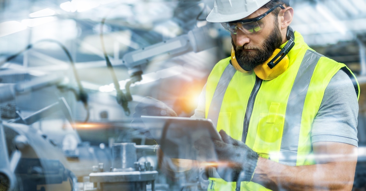 A man in a hard hat and hi-vis vest standing among machinery while looking at a tablet in his hands.