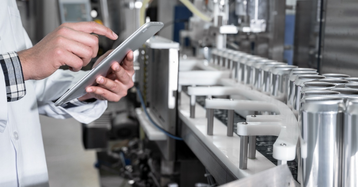 A person wearing a white coat and holding a tablet stands next to a conveyor full of shiny metal cylinders.
