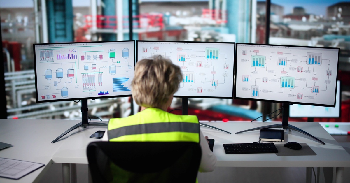 A person in a hi-vis vest sitting at a desk with three large monitors displaying data, charts, and graphics.